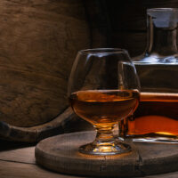 Glass cognac, bottle of cognac and an old oak barrel of cognac with blurred background in the light rays. Table on a wooden stand there is decanter of skate and glass of cognac against.